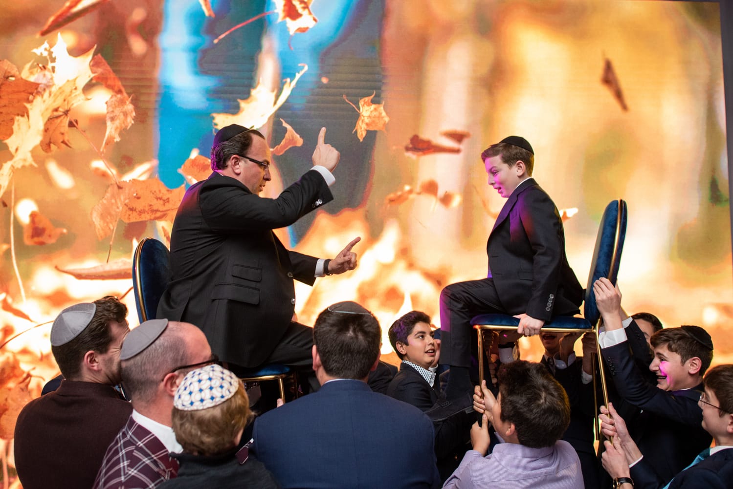 Bar mitzva boy with his dad being lifted up on chairs by men in suits at a party in front of an orange background with leaves